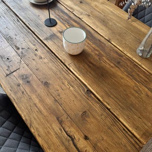 Könnte beinhalten: Ein rustikaler Esstisch aus Holz mit warmer, natürlicher Oberfläche. Der Tisch besteht aus breiten Brettern und weist sichtbare Holzmaserung und Knoten auf. Eine weiße Keramikschale mit gelbem Muster und ein schwarzer Kerzenhalter befinden sich auf dem Tisch.