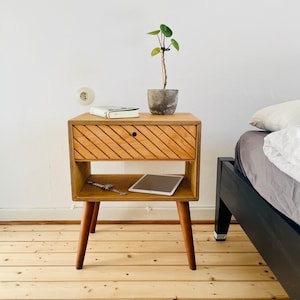 Könnte beinhalten: Ein Nachttisch aus Holz mit einer Schublade mit diagonalen Rillen und einem unteren Regal. Der Nachttisch hat konische Beine und beherbergt eine kleine Topfpflanze, ein Buch, eine Uhr und ein Tablet. Das Holz hat eine warme, natürliche Oberfläche.
