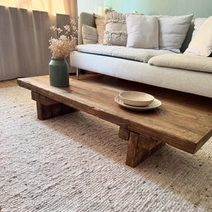 Könnte beinhalten: Ein rustikaler Holz-Couchtisch mit natürlicher Holzoptik und schlichtem Design. Der Tisch hat zwei dicke, rechteckige Beine und eine breite, flache Tischplatte. Eine weiße Keramikschüssel steht auf dem Tisch, und eine Vase mit getrockneten Blumen steht auf dem Boden neben dem Tisch.