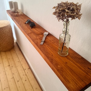 Könnte beinhalten: Ein braunes Holzregal an einer weißen Wand. Es zeigt eine Sonnenbrille, eine silberne Armbanduhr, eine klare Glasflasche mit getrockneten Blumen und zwei weiße Keramikvasen. Das Regal ist etwa 120 cm lang.