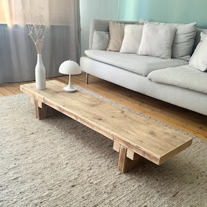 Op de afbeelding: Een lichte houten salontafel met een rechthoekig blad en twee sets poten. De tafel staat op een lichtbeige kleed voor een lichtgrijze bank met kussens.