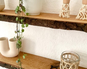 Puede incluir: Dos estantes de madera rústica con borde natural. El estante superior contiene una maceta blanca acanalada con una planta colgante. El estante inferior muestra un jarrón crema, un portavelas de vidrio con detalles tejidos y una planta colgante.