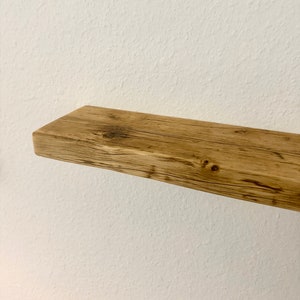 Könnte beinhalten: Rustikales Holzregal mit natürlicher, verwitterter Optik. Das Regal besteht aus hellbraunem Holz mit sichtbarer Maserung und Knoten und ist an einer weißen Wand befestigt. Das Regal ist etwa 76 cm lang.