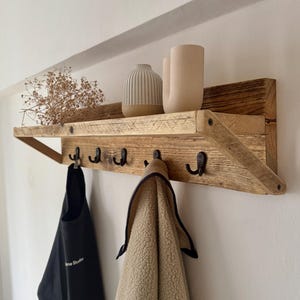 May include: A rustic wooden shelf with five hooks and a small shelf on top. The shelf is made of reclaimed wood and has a natural finish. There are two ceramic vases on the top shelf, one with a white and gray striped pattern and the other with a solid beige color. A black apron and a beige fleece jacket are hanging on the hooks.