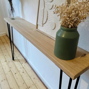 Può includere: Un tavolo console in legno con gambe a forcina nere. Il tavolo è decorato con una stampa d'arte astratta incorniciata bianca e un vaso verde con fiori secchi.