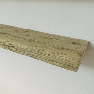 Könnte beinhalten: Ein rustikales, verwittertes Holzregal, montiert an einer weißen Wand. Das Regal hat eine helle, natürliche Holzfarbe mit sichtbarer Maserung und Knoten, was auf eine wiederverwertete oder abgenutzte Ästhetik hindeutet. Das Regal ist rechteckig und scheint zur Präsentation bereit zu sein.