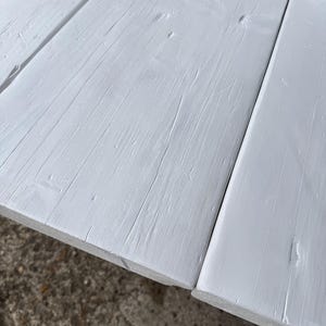 Könnte beinhalten: Eine Reihe paralleler Holzplanken mit matter weißer Lackierung. Die Oberfläche der Bretter zeigt eine sichtbare Holzmaserung mit natürlichen Vertiefungen, Astlöchern und linearen Rillen, die entlang der Länge jedes einzelnen Brettes verlaufen.