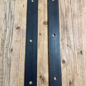 Puede incluir: Dos tiras de metal negro con agujeros perforados a lo largo de su longitud. Las tiras están sobre una superficie de madera.