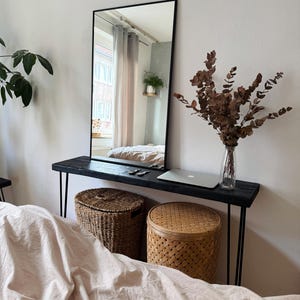 Può includere: Un grande specchio nero a figura intera riflette una camera da letto. Un tavolo consolle in legno scuro con gambe in metallo nero sostiene un laptop e un vaso con rami secchi. Due cesti intrecciati sono posizionati sotto.