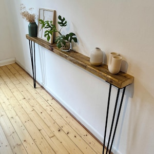 May include: A rustic wooden shelf with black hairpin legs, decorated with a small potted plant, a framed print, and three ceramic vases. The shelf is mounted on a white wall above a wooden floor.