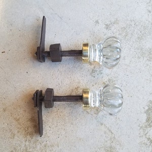 Op de afbeelding: Twee antieke glazen deurknoppen met metalen stelen en een metalen hendel. De knoppen zijn van helder glas met een getextureerd ontwerp. De stelen zijn van bruin metaal met een gouden accent.