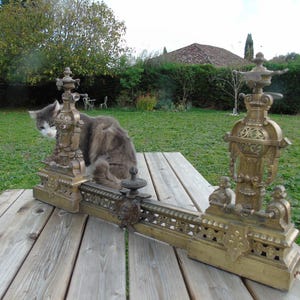 Op de afbeelding: Oud messing haardstel met sierlijke krullen en decoratieve ornamenten. Het haardstel staat buiten op een houten tafel. Het messing heeft een warme, verouderde patina. Een kat zit in de buurt.