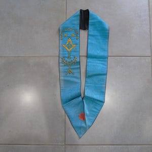 Peut inclure: Une écharpe cérémonielle bleu clair ornée de symboles brodés en fil doré, incluant une équerre et un compas, des motifs floraux et une petite lettre A en bas. L'écharpe présente une extrémité pointue avec un emblème rouge et une doublure noire.