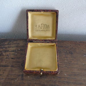 Pode incluir: Uma caixa de joias quadrada com dobradiças e acabamento exterior salpicado em castanho e preto. O interior é forrado com tecido dourado, apresentando o texto SARDA BESANÇON impresso em tinta escura no interior da tampa. Um pequeno fecho é visível.
