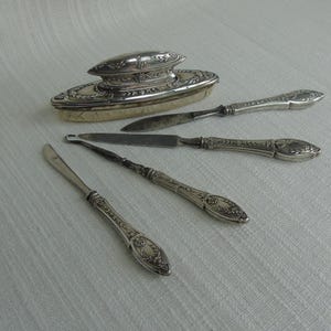 May include: An antique silver vanity set, including a powder box and several small knives or tools. The set features ornate floral detailing and a classic design. The items are arranged on a white, textured surface.