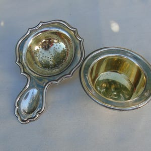 May include: A silver tea strainer and drip bowl. The strainer has a handle and a perforated bowl with a decorative scalloped edge. The drip bowl is round with a silver rim. The items are likely vintage, suitable for tea.