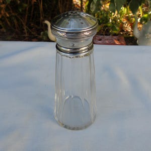 May include: A clear glass salt shaker with a silver metal top. The shaker has a ribbed design on the glass body and a perforated top for dispensing salt. The salt shaker is a vintage kitchenware item.