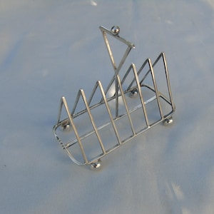 Op de afbeelding: Een zilverkleurige toastrek met een geometrisch ontwerp. De rek heeft driehoekige verdelers en rust op vier kleine, afgeronde voetjes. Een driehoekige handgreep met een bolvormige bekroning komt van bovenaf.