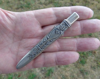 Op de afbeelding: Een zilverkleurig metalen mesheft met een gedetailleerd reliëfpatroon van bloemen en bladeren langs de schacht. Het heft heeft een gladde, afgeronde punt en een cilindervormig bovendeel met horizontale ribbels en een metalen afwerking.