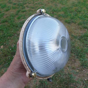 Puede incluir: Una lámpara de exterior redonda y vintage con una lente de vidrio transparente y acanalada. La lámpara tiene un marco de metal color crema con detalles en color latón. La luz es sostenida por una mano sobre un fondo de hierba verde.