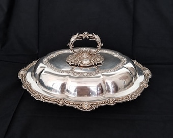 Può includere: Un piatto da portata ovale in argento con coperchio a cupola e manico decorativo. L'oggetto presenta complessi rilievi lungo i bordi smerlati e una base del manico ornata, caratterizzato da una finitura metallica lucida su tutta la superficie.