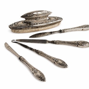 Könnte beinhalten: Ein Set aus drei silberfarbenen Metallwerkzeugen mit verzierten, geprägten Griffen mit Blatt- und Muschelmotiven, begleitet von einem ovalen silberfarbenen Schreibtischaccessoire mit passendem geprägtem Deckel und einer strukturierten Basis.