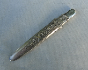 Op de afbeelding: Een zilveren briefopener met sierlijke bloemen- en geometrische ontwerpen. De opener heeft een rechthoekige handgreep en een puntig lemmet. Het metaal heeft een gepolijste afwerking, die licht reflecteert. Het ontwerp is elegant en gedetailleerd.