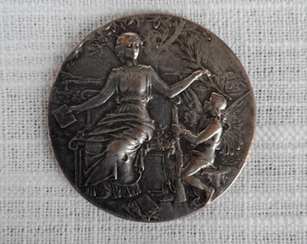 Puede incluir: Una medalla redonda plateada con un diseño en relieve de una mujer sosteniendo un bastón y una figura alada arrodillada ante ella.