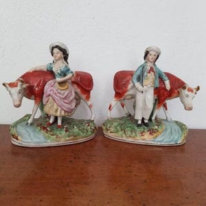 Puede incluir: Dos figuras de cerámica que representan a un niño y una niña con vacas. Las vacas son rojas y blancas, y las figuras visten ropa de época. Las figuras están sobre una base con hierba pintada y un arroyo.