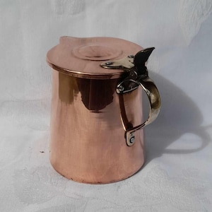 Puede incluir: Una jarra de cobre con tapa y asa de latón. La jarra tiene un acabado brillante y pulido.
