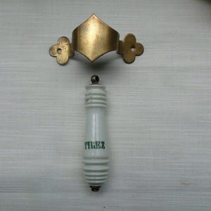 Op de afbeelding: Witte porseleinen toiletgreep met groene tekst "TIREZ". De greep is bevestigd aan een gouden metalen beugel met een ruitvormige bovenkant en decoratieve zijstukken.