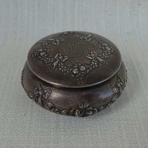 Può includere: Un'antica scatola portagioie rotonda color argento. Il coperchio e la base sono adornati da disegni floreali e a nastro. Il coperchio presenta un monogramma inciso all'interno di una ghirlanda floreale. La scatola ha un aspetto ossidato e invecchiato.