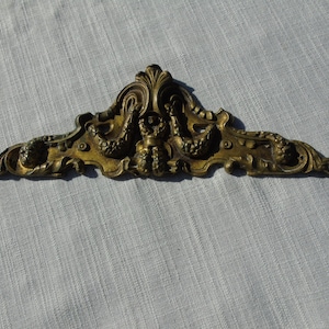 Può includere: Un'ornamento decorativo in metallo dorato con un design floreale stilizzato. L'ornamento ha un motivo centrale di un viso stilizzato con una corona e un paio di ali.