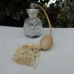 Puede incluir: Un frasco de perfume vintage con un cuerpo de vidrio transparente tallado en forma de diamante y una tapa de metal plateado. Cuenta con un atomizador de bulbo beige con un tubo largo y curvo y una borla. El frasco está sobre una superficie blanca.