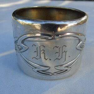 Op de afbeelding: Een zilveren servetring met gegraveerde initialen "I.F." en decoratieve Art Nouveau-stijl versieringen. De cilindrische ring heeft een gepolijst oppervlak dat het licht reflecteert. De servetring is een vintage item.