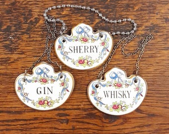 May include: Three porcelain bottle labels with gold trim and floral designs. The labels are attached to silver chains and read "Gin", "Sherry", and "Whisky".