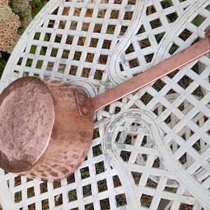 May include: A copper saucepan with a long handle and a hammered texture. The pan is resting on a white, lattice-patterned table. The handle has a loop for hanging. Pink flowers and green foliage are in the background.