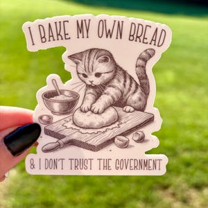 Puede incluir: Una pegatina con un gato amasando masa en una tabla de madera, con un cuenco y un rodillo. El texto dice "I BAKE MY OWN BREAD & I DON'T TRUST THE GOVERNMENT". El gato es marrón y blanco, sobre un fondo verde.