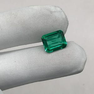 Puede incluir: Una esmeralda verde vibrante, tallada en forma rectangular. La gema se muestra sobre un fondo de tela blanca, resaltando su claridad y color. Las facetas de la esmeralda reflejan la luz, destacando su brillo. Ideal para joyería.