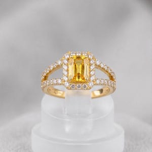 Könnte beinhalten: Ein goldener Ring mit einem rechteckigen gelben Edelstein, umgeben von einem Halo aus kleineren weißen Edelsteinen. Der Ring hat ein geteiltes Schaftdesign mit zusätzlichen weißen Edelsteinen entlang des Bandes. Der Ring wird auf einem transparenten Ständer präsentiert.