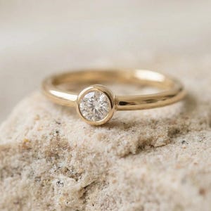 Könnte beinhalten: Ein goldener Ring mit einem runden, klaren Diamanten in einer Lünettenfassung. Der Ring liegt auf einer hellen, strukturierten Oberfläche. Das Band ist glatt und poliert.