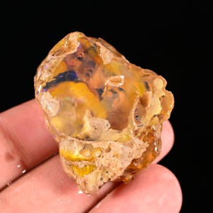 Könnte beinhalten: Ein roher, ungeschliffener Opal-Edelstein mit einer Mischung aus Farben. Der Opal hat eine Basis aus Beige und Braun mit leuchtenden Blitzen in Gelb, Orange und Blau. Der Stein hat eine natürliche, unregelmäßige Form und eine raue Textur.