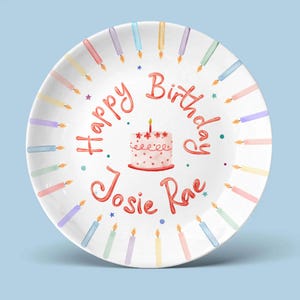 May include: White ceramic plate with a birthday theme. The plate features the words "Happy Birthday Josie Rae" in red script, a small cake, and colorful candles around the rim. The background is a light blue.