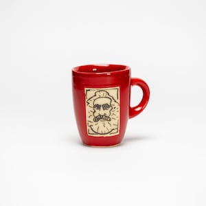 Könnte beinhalten: Eine rote Keramiktasse mit einer handgezeichneten Illustration eines Mannes mit Schnurrbart und Bart. Die Tasse hat ein schlichtes Design mit einem abgerundeten Körper und einem Henkel.