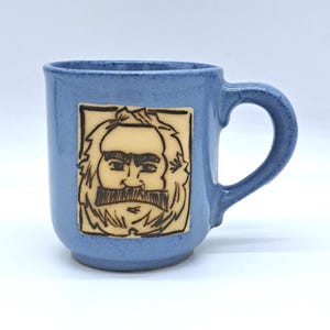 Könnte beinhalten: Eine blaue Keramiktasse mit einem handgezeichneten Bild eines Mannes mit Bart und Schnurrbart. Das Bild befindet sich in einem quadratischen Rahmen und ist in hellbrauner Farbe.