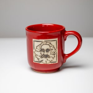 Könnte beinhalten: Eine rote Keramiktasse mit einem weißen quadratischen Design, das eine Schwarzweißillustration eines Mannes mit Bart zeigt.