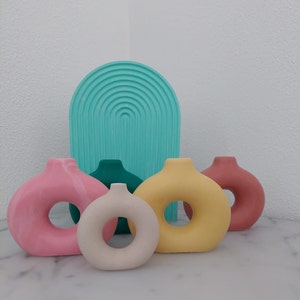 Könnte beinhalten: Eine Sammlung farbenfroher, moderner Dekorationsvasen. Die Vasen sind in den Farbtönen Rosa, Gelb, Türkis, Grün und Beige gehalten. Die Vasen haben eine einzigartige Donut-Form mit einem Loch in der Mitte. Ein türkis geformtes Dekorationselement befindet sich hinter den Vasen.
