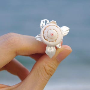 May include: A silver ring with a seashell design. The ring features a spiral shell with a pink centre, set within silver detailing. The ring is held against a blurred blue backdrop, possibly a beach or ocean scene.