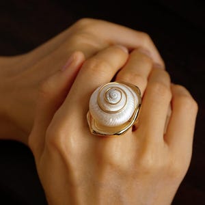 May include: A ring featuring a white, spiraled seashell centrepiece mounted on a polished gold-toned metal band. The shell displays a natural, coiled texture with a pearlescent sheen, securely set within a smooth, rounded gold-coloured frame.