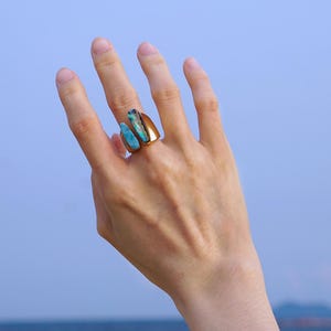 May include: A gold ring with three turquoise stone features. The ring is worn on a hand, with a blue sky and sea in the background. The ring's design is modern, highlighting the natural beauty of the stones. The ring is a unique piece of jewellery.